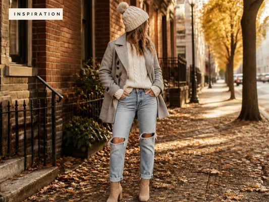Woman on a fall city sidewalk wearing a light grey wool peacoat, cream cable-knit sweater, ripped boyfriend jeans, tan suede ankle booties and a cream knit beanie.