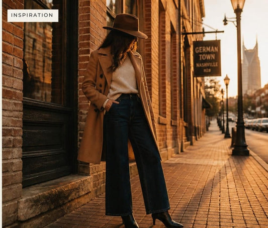 Woman on a Germantown Nashville sidewalk wearing a long camel pea coat, cream crewneck sweater, dark wide-leg jeans, black kitten-heel ankle boots and a brown wide-brim fedora.